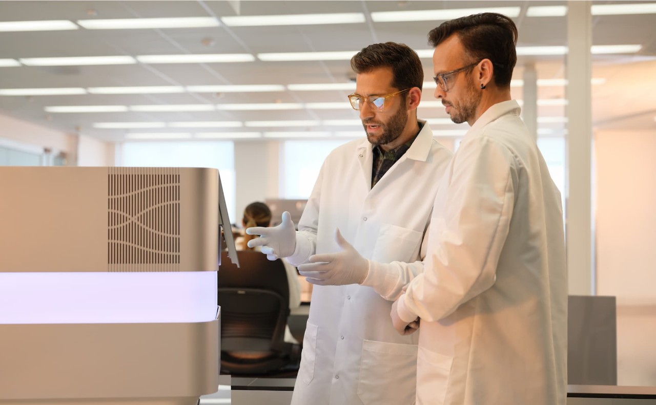 Profile image of two male scientists, one training, looking at the start screen on a NextSeq 1000/2000 in a dry lab; female scientist sitting at a desk in the background.