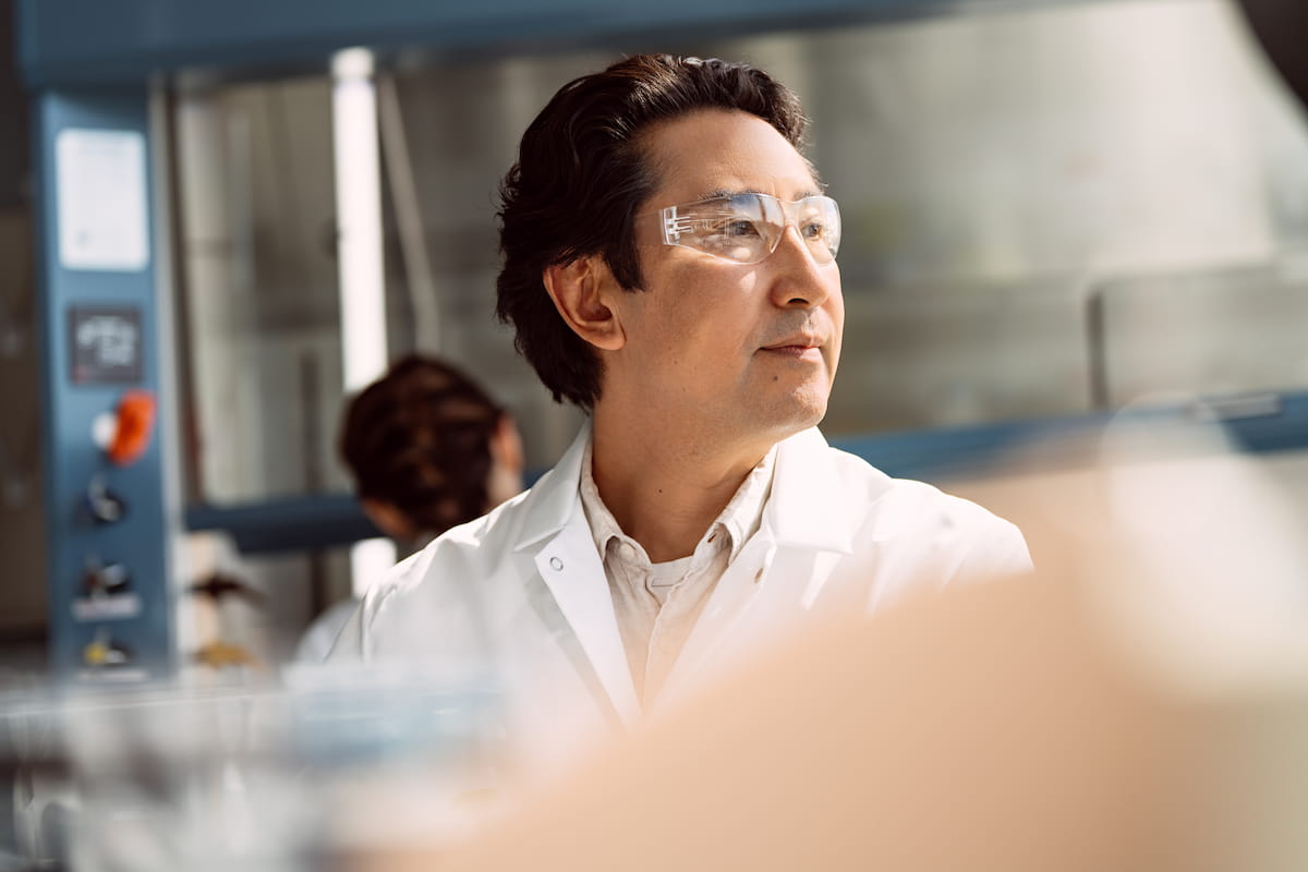 Male scientist looking at something, not visible, in a lab setting; blurry image of a scientist in the background.
